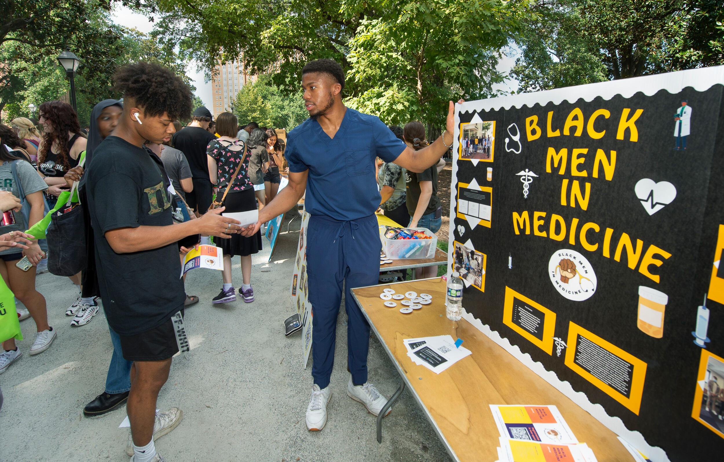 A medical student hands another student materials at the Black Men in Medicine booth of the Student Organization & Volunteer Opportunities Fair this fall along a sidewalk next to a tree-lined green space in Monroe Park.