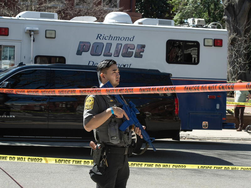 A photo of a man in a police uniform holding a fake blue gun and standing. Around him is police caution tape and behind him is a black SUV and a white and blue van that reads \"Richmond POLICE\"