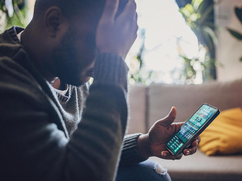 A photo of a man holding his head in his hand while looking at a sports betting app on his phone.