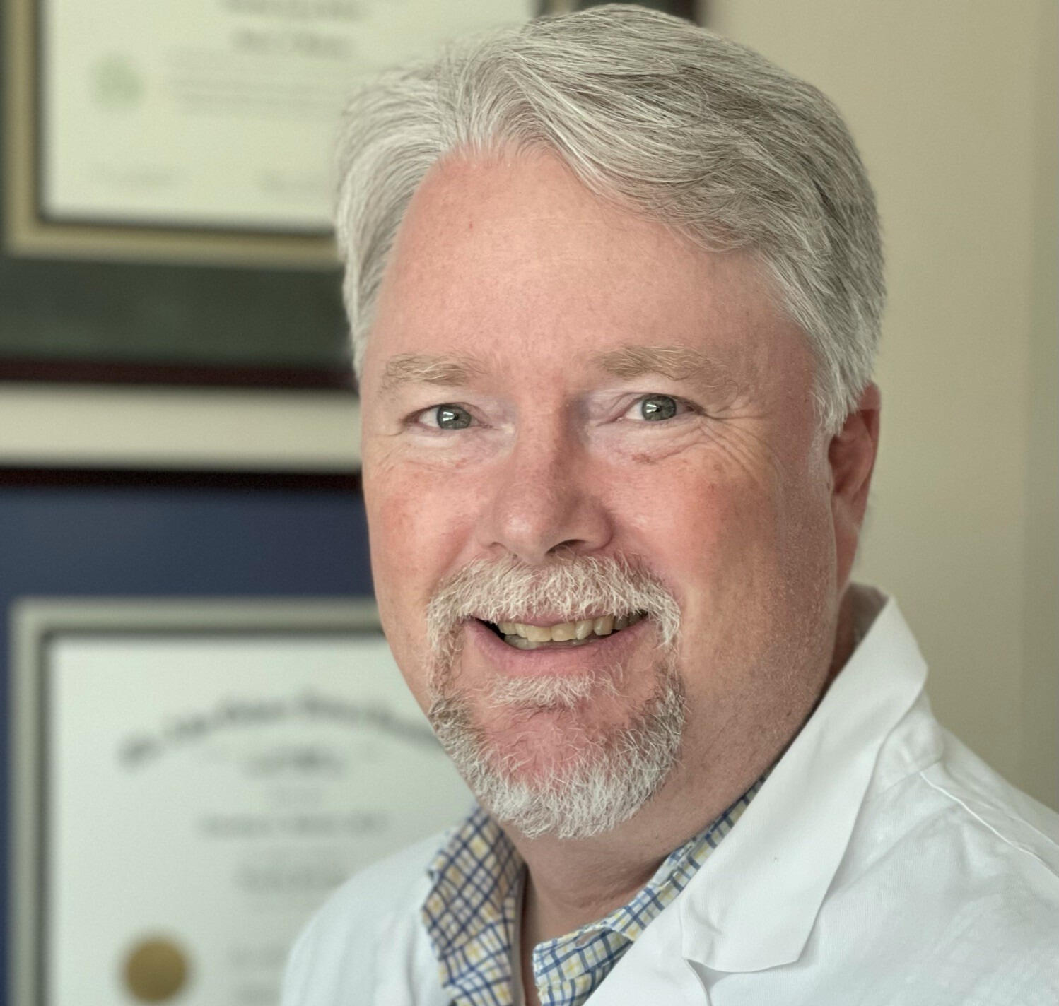 A photo of a man wearing a white medical coat. On the wall behind him are two framed degrees. 