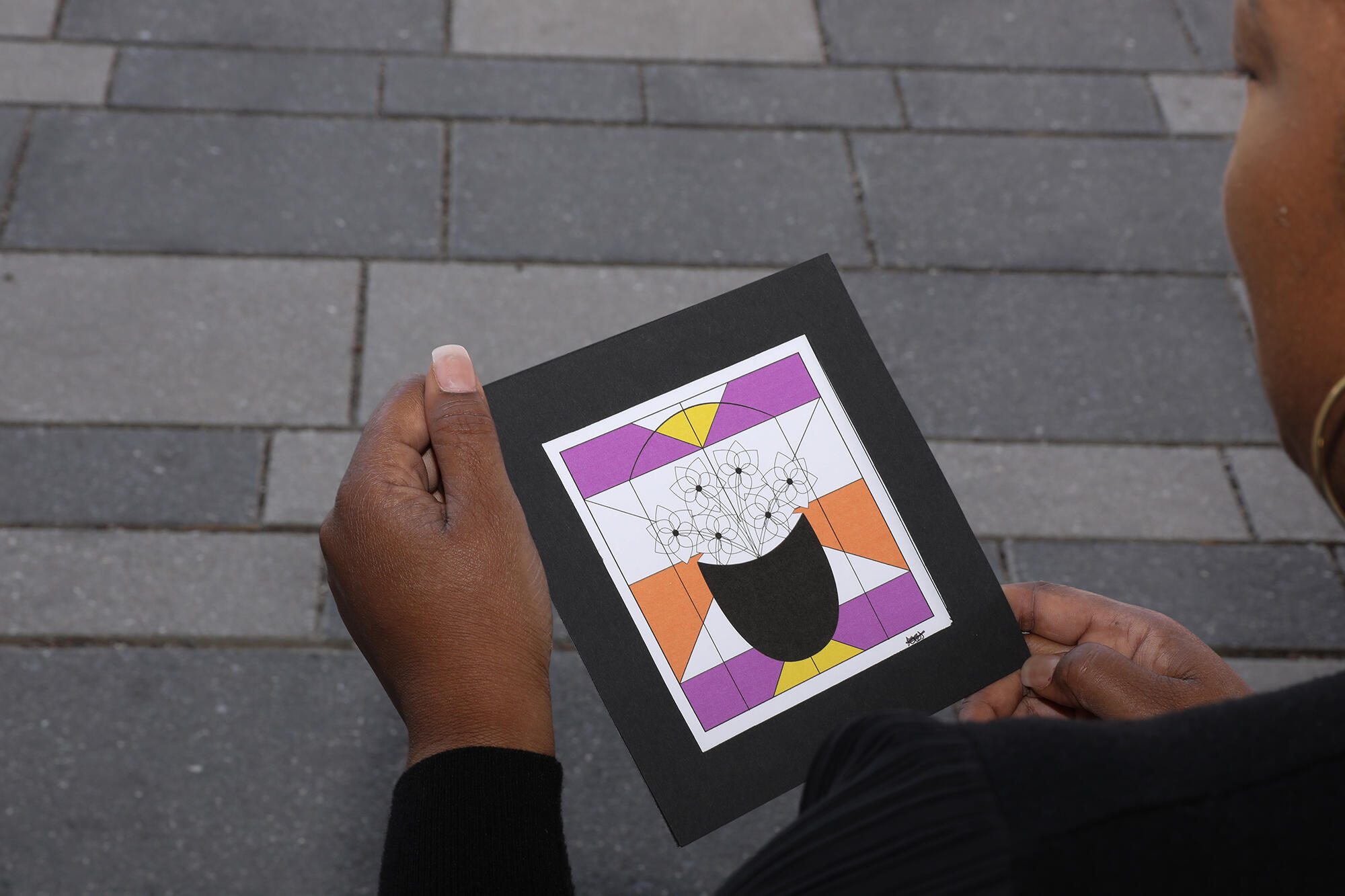 A photo of a woman from behind. She is holding an illustration of a pot of flowers with geometric shapes in the background of the picture.