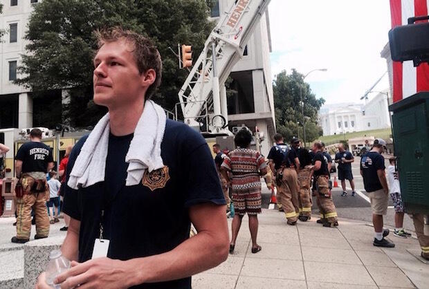 A man holding a plastic bottle with a towel wrapped around his neck in the foreground. Firefighters.