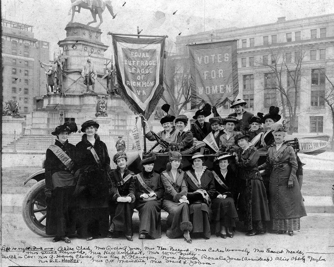 A group of women is posed in front of the Virginia Washington Monument on the public square in Richmond, Virginia. Two members are holding poles with banners affixed atop them. One banner says \"Equal Suffrage League of Richmond, VA,\" the other says \"Votes For Women.\" The bottom of the photo is inscribed with a list of those in the photo.