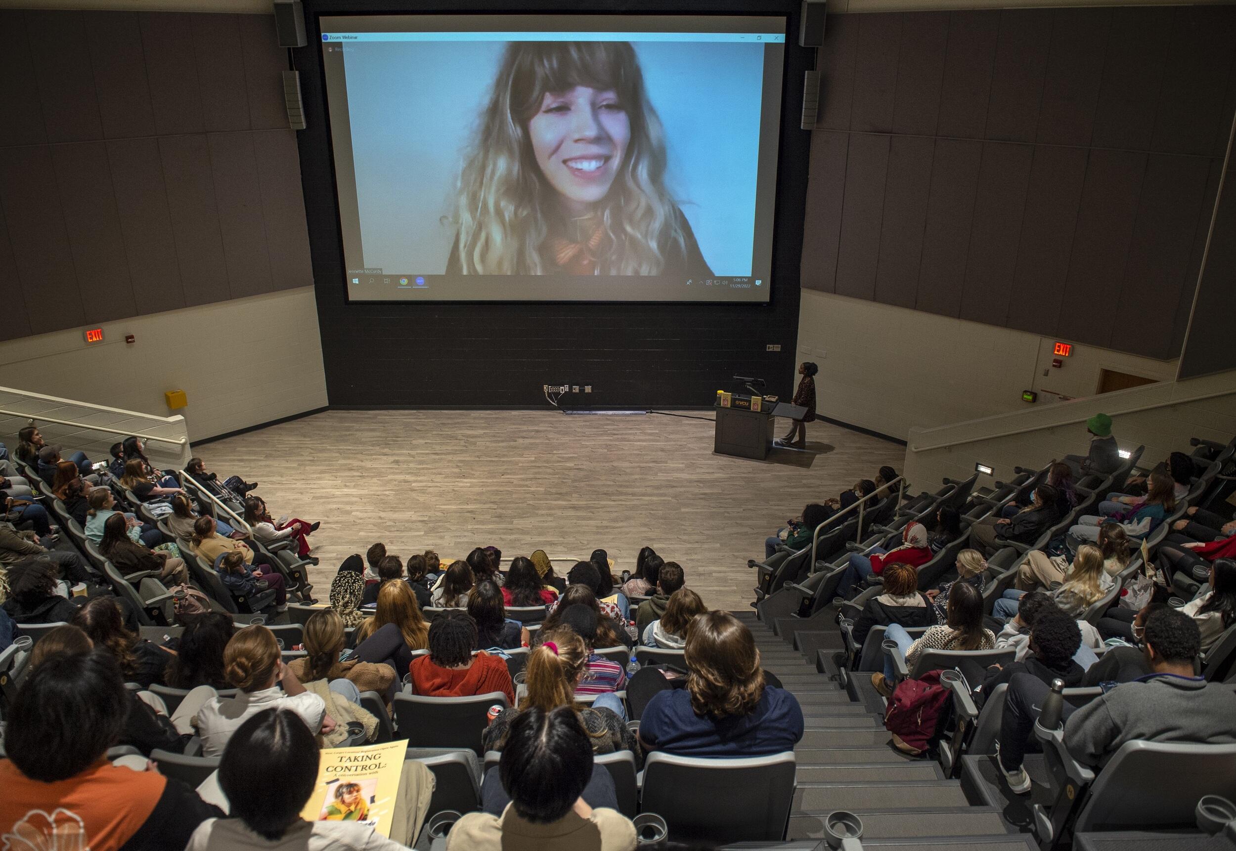 An auditorium of students facing a projector with Jenette McCurdy on it. 