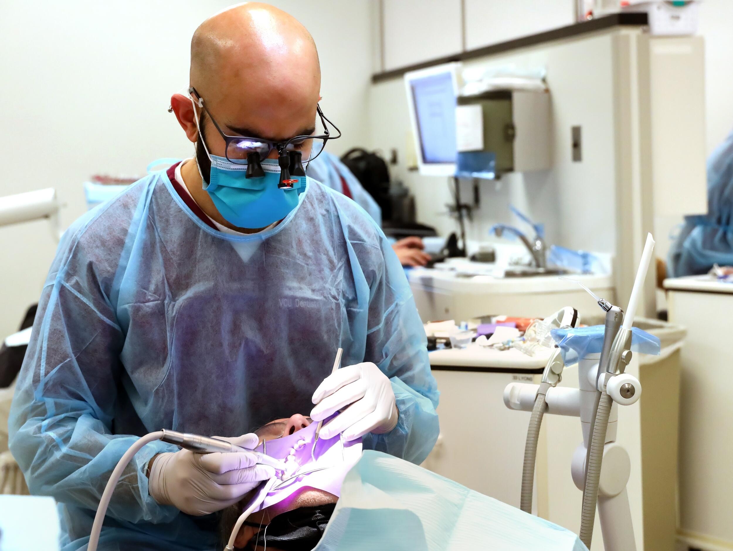 A photo of a man wearing blue scrubs and a face mask cleaning the teeth of another person sitting in a dental chair in front of him. 