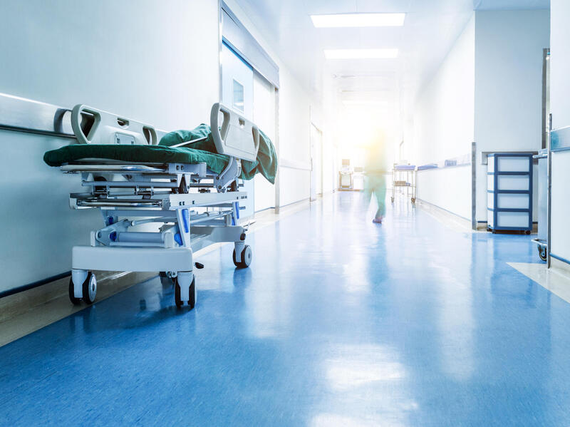A photo of the hallway of a hospital with a stretcher sitting against the left side wall. Down the hallways is a figure walking who is distorted by a beam of light at the end of the hall. 