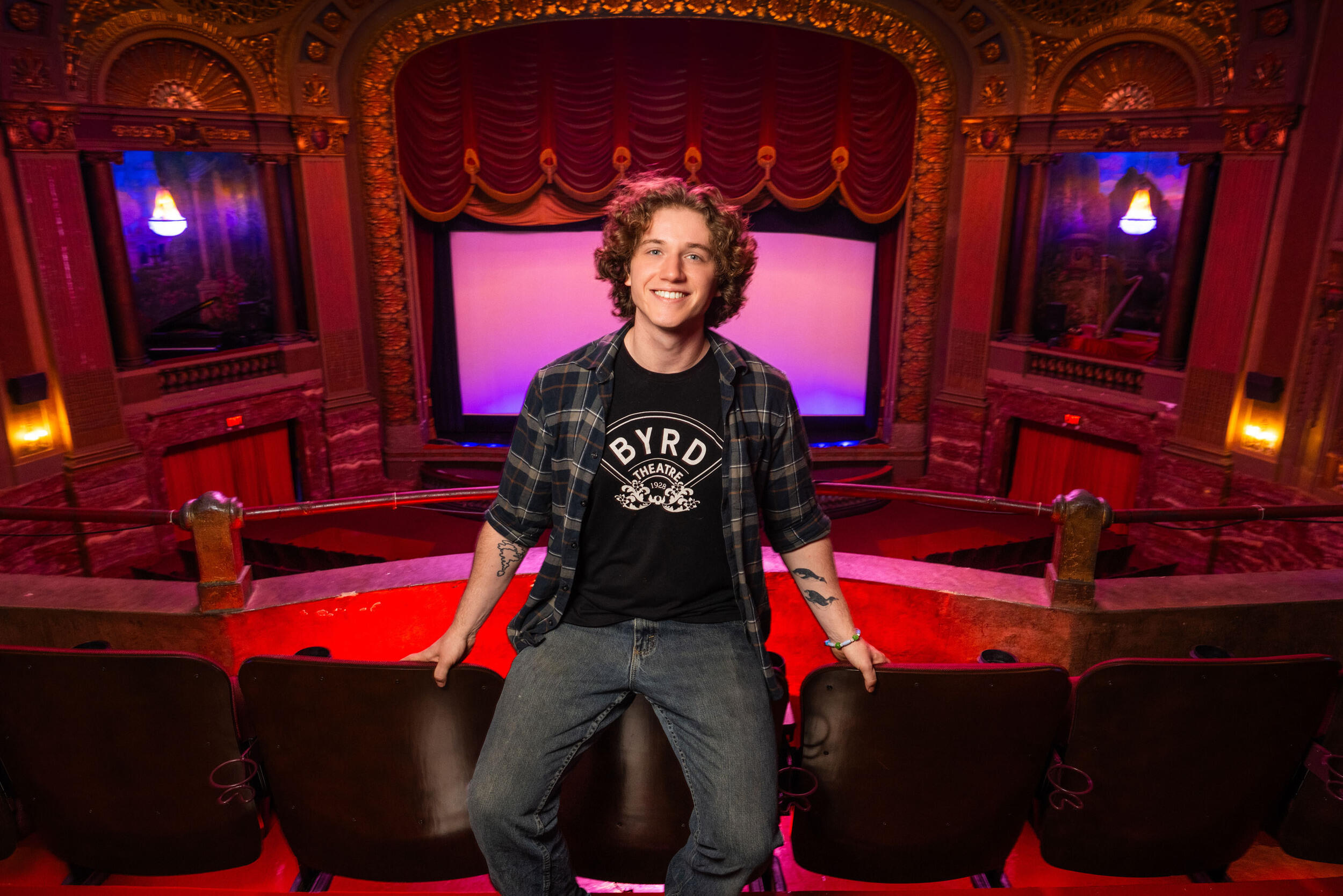 A photo of a man sitting in an ornate, old-fashioned movie theater. The screen where the movies are shown has a red velvet curtain. The man is wearing a back T-shirt that says \"BYRD\" in white letters. 