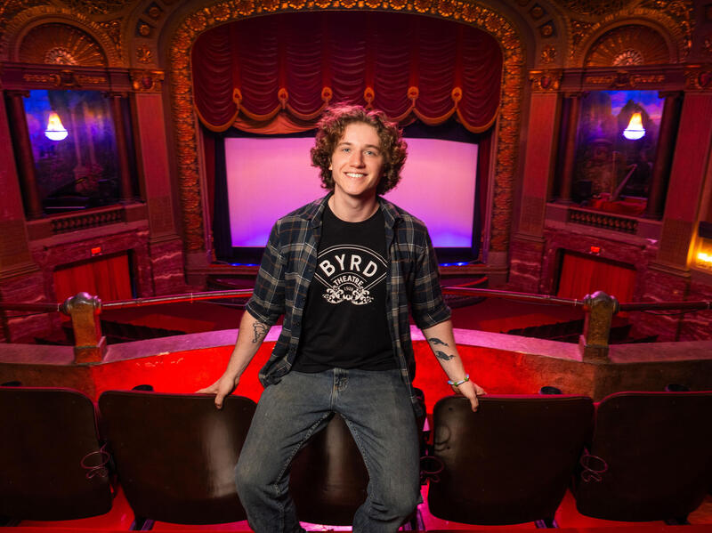 A photo of a man sitting in an ornate, old-fashioned movie theater. The screen where the movies are shown has a red velvet curtain. The man is wearing a back T-shirt that says \"BYRD\" in white letters. 