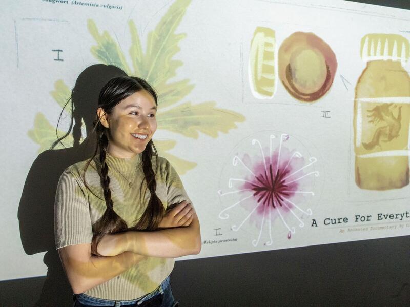 A photo of a woman standing against a wall with her arms crossed over her chest. A projection is being displayed on the wall. It has the image of a leaf, a circular plant, and a bottle with a screw top from the top and side. Black text at the bottom right of the projection says \"A Cure For Everything\"