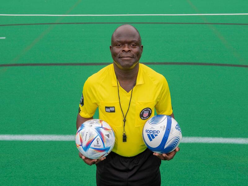 Kwasi Deitutu, wearing a soccer ref's uniform, holds two soccer balls.