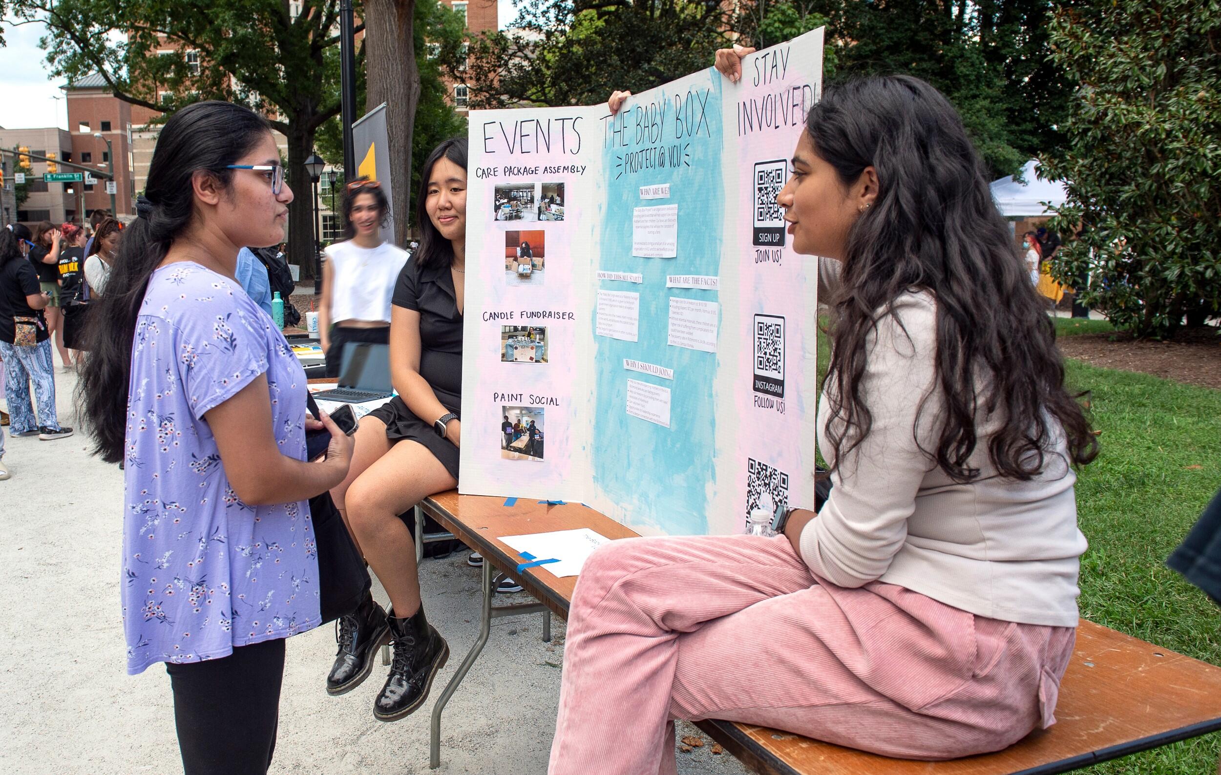 Two students seated on a table next to a poster that says The Baby Box Project at VCU talk with another student on a sidewalk next to a tree-lined green space in Monroe Park.