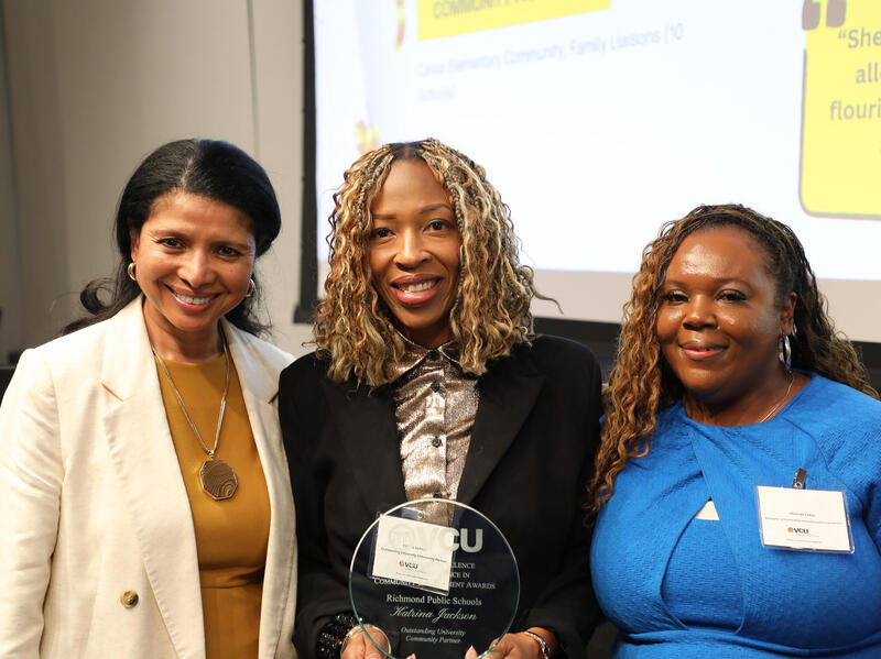 A photo of three woman from the chest up. The woman in the center is holding a glass award trophy. 