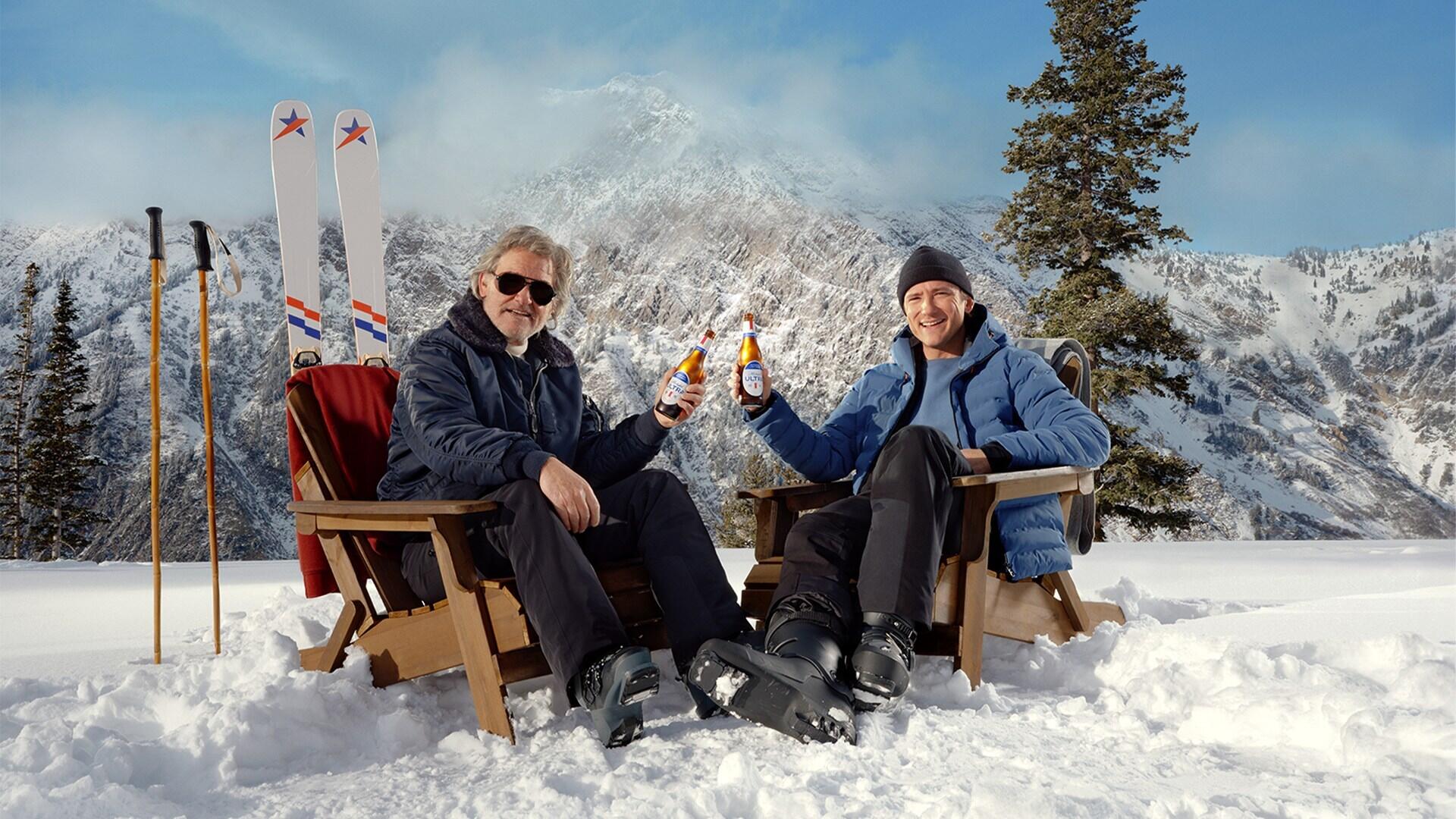 A photo of two men sitting on a snowy moutain in wooden chairs. Each man is holding a Michelob ULTRA beer. Behind the man on the left is a pair of skis. 