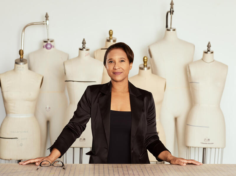 A photo of a woman from the waist up standing behind a table. Behind the woman are seven sewing mannequins.