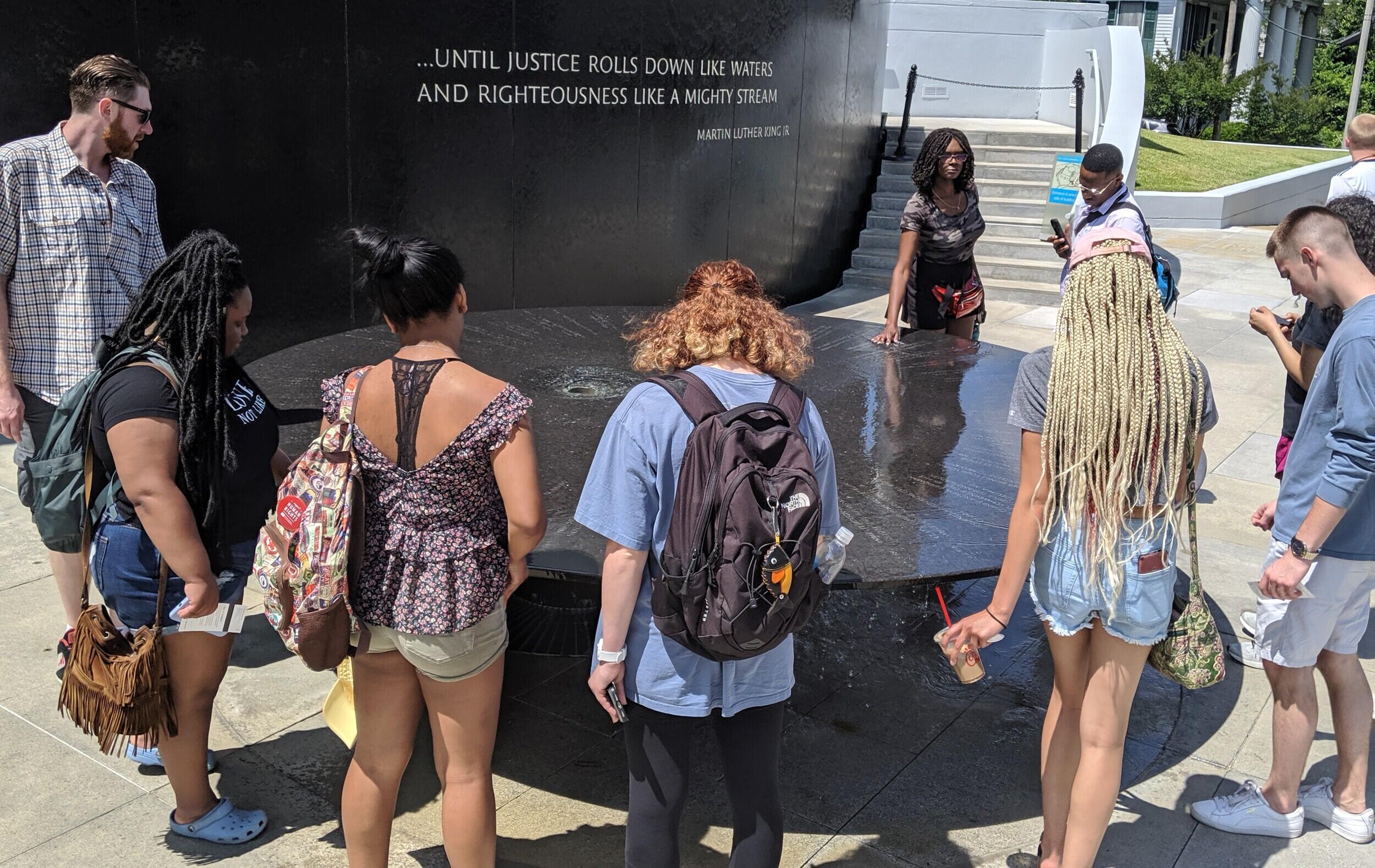 A group of students surrounding a circular granite monument. A quote is listed on the wall behind the monument.