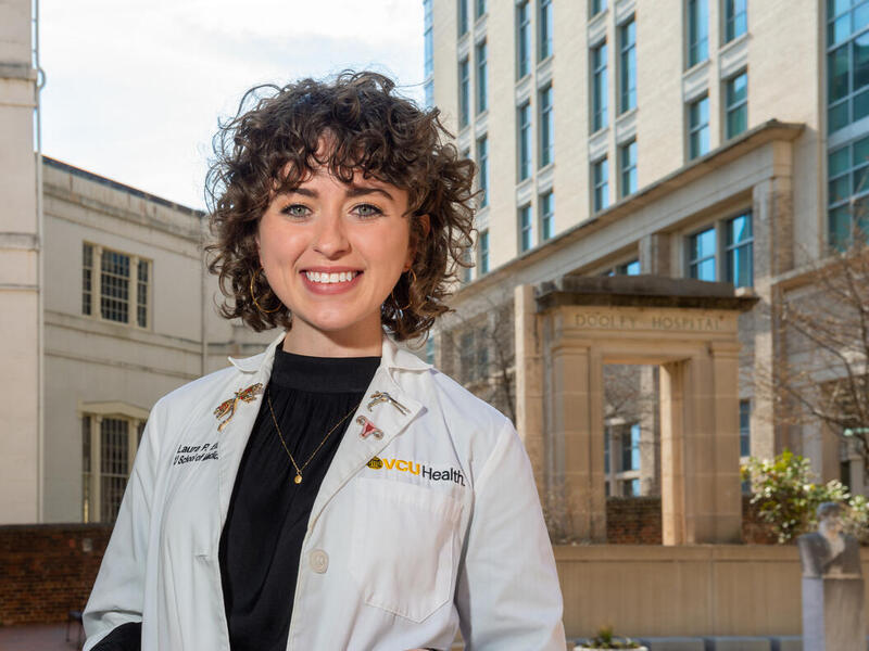 Laura Ellis smiling and wearing a white lab coat that says \"VCU Health\" 