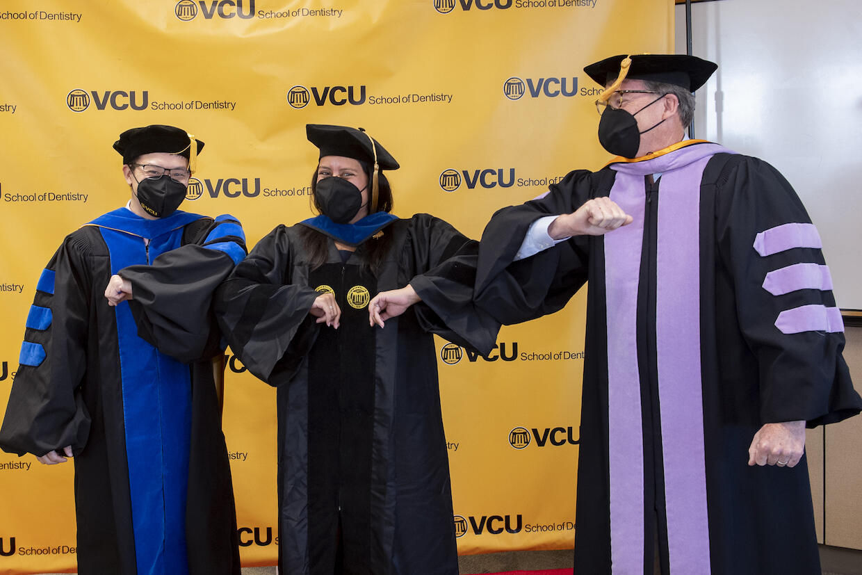 Three people in cap and gown wearing masks. A V C U School of Dentistry backdrop sits behind them.