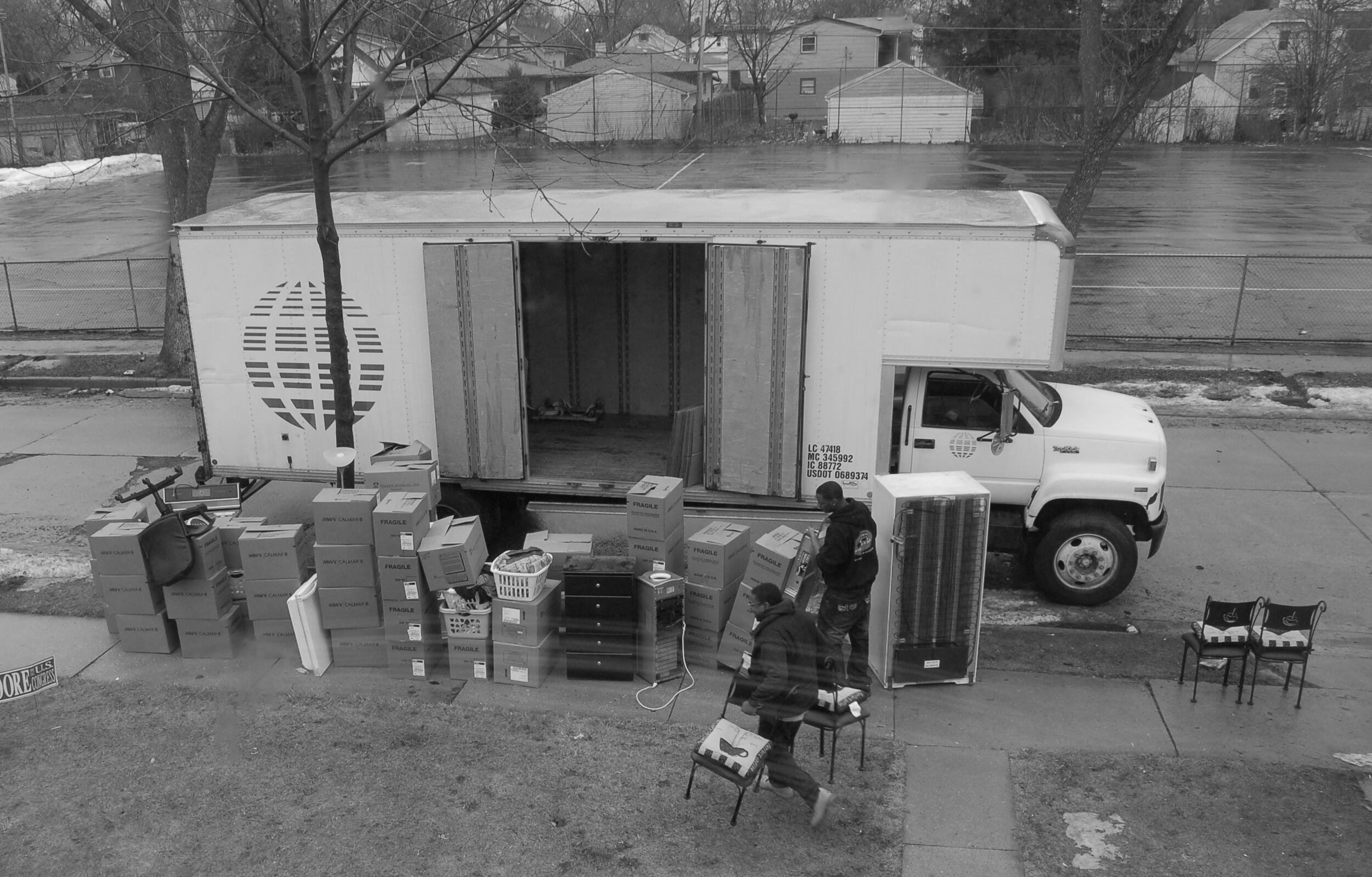 A moving van with its side door open is parked on a street. Various household items and moving boxes sit alongside it. Two moving assistants are putting items next to the van.