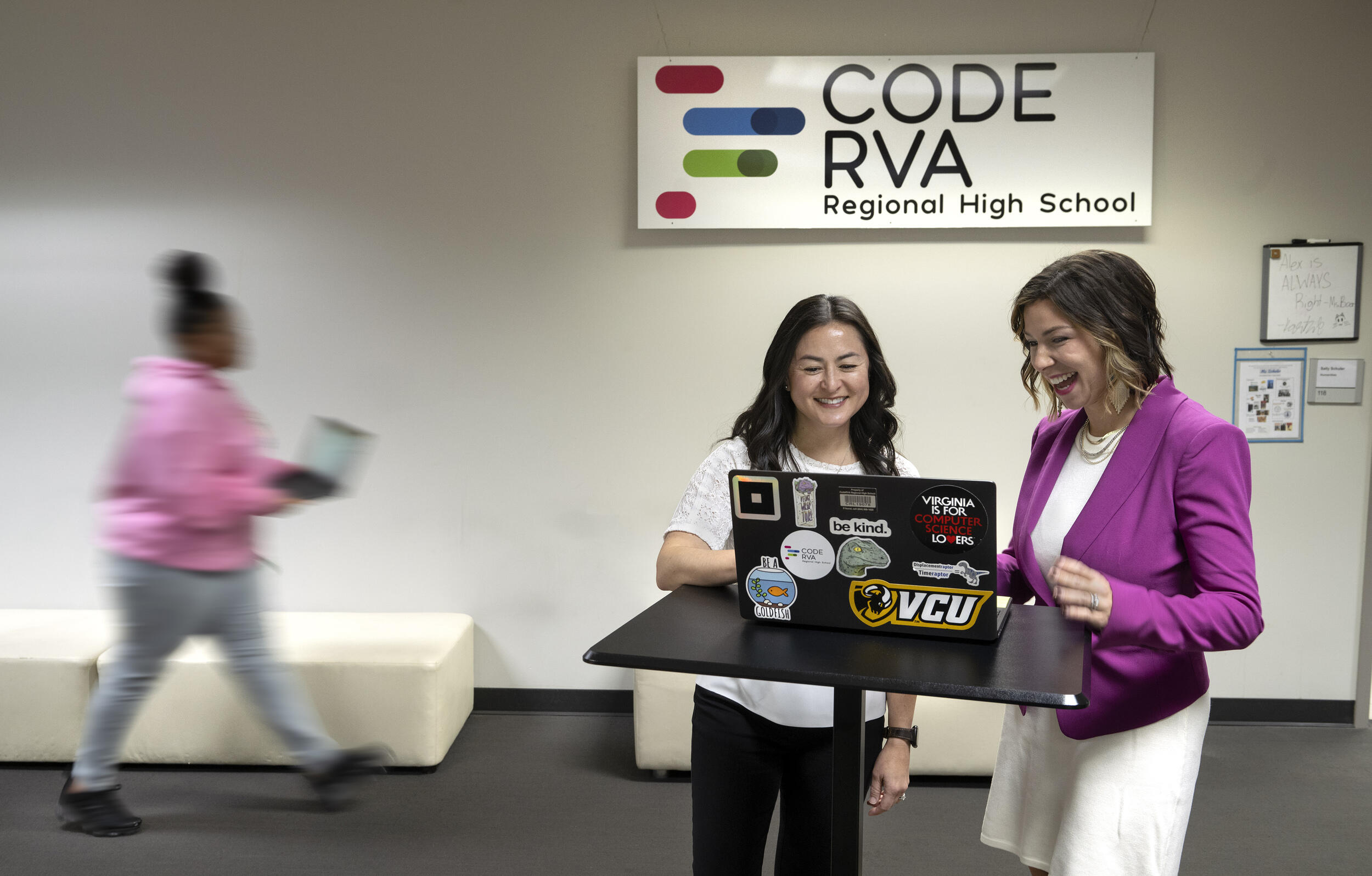 A photo of two women looking at a laptop on a table. Behind them is a sign that reads \"CODE RVA Regional High School.\"