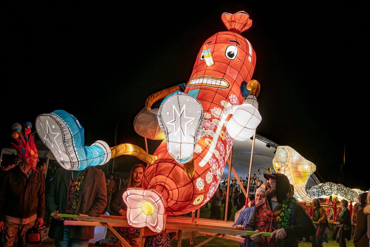 A photo of a large paper sculpture of a hot dog with arms, leggs, and a face. 