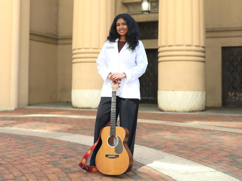 A woman in a white coat stands and holds a guitar.