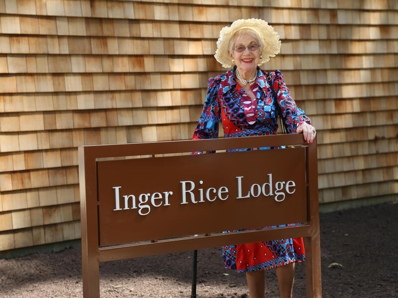 A photo of a woman smiling behind a brown sign that says \"Inger Rice Lodge\" in white letters. 