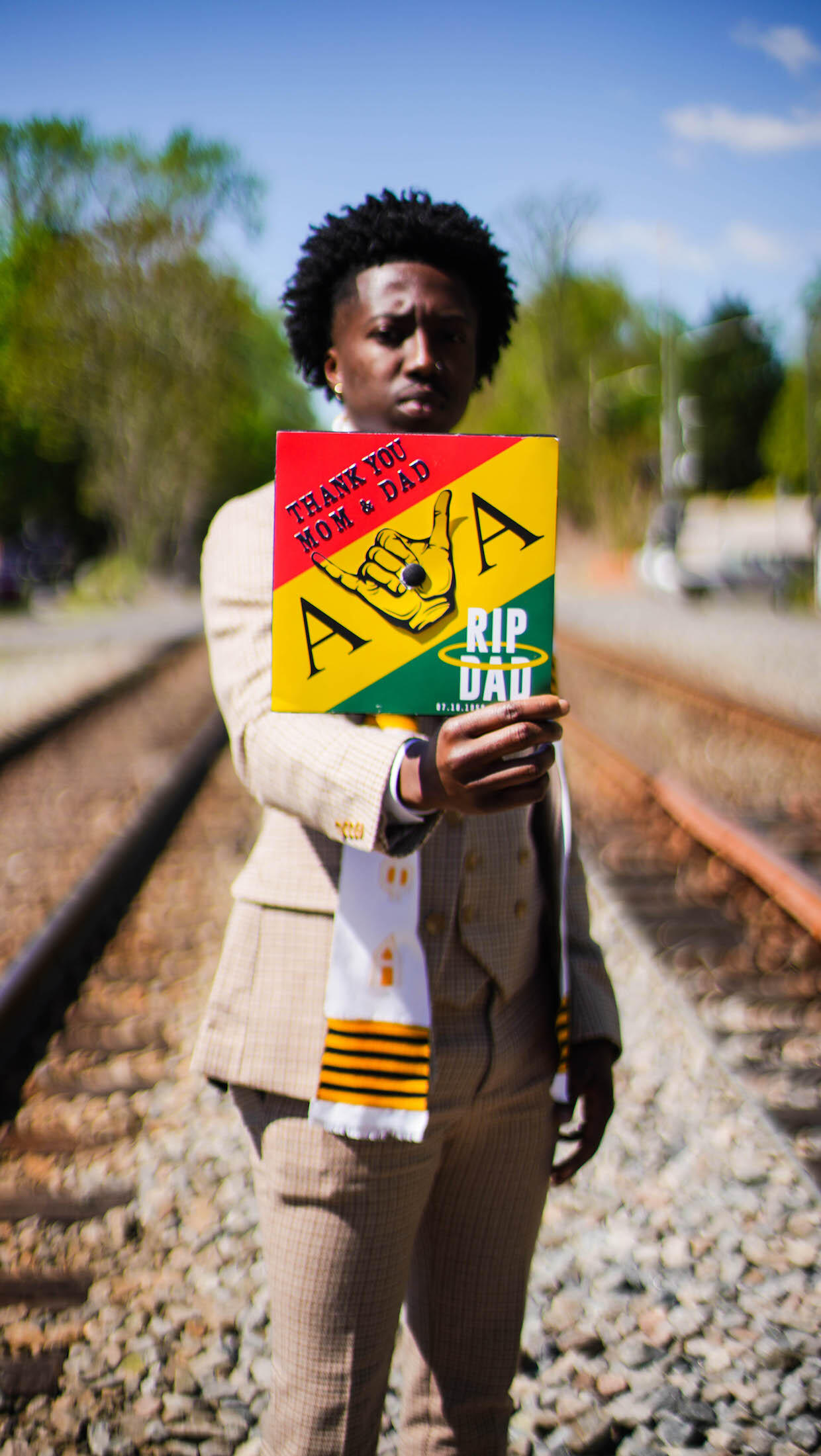 Gideon Boakye standing on train tracks, holding his graduation mortarboard, which is decorated with messages to his parents laid over the red, yellow and green stripes of the Ghanaian flag.