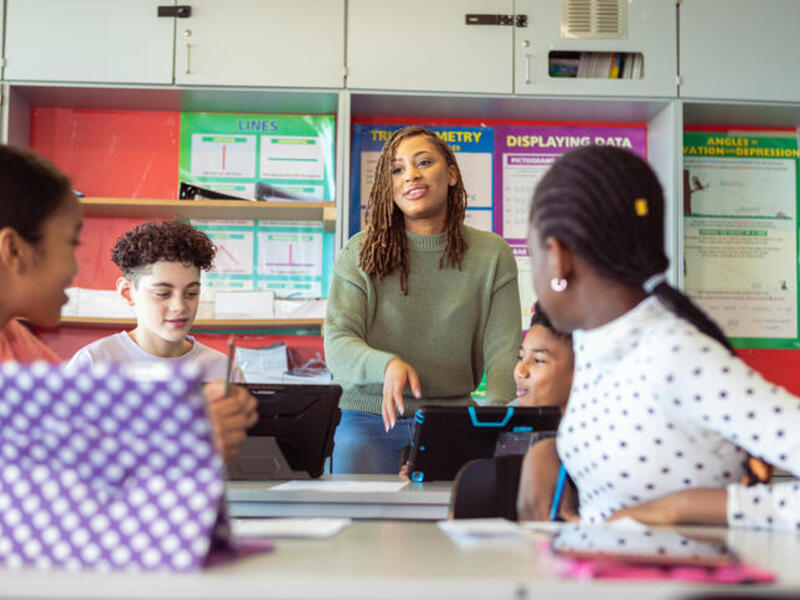 A photo of a teacher talking to four students sitting at two tables in front of her. The two boys are holding tablets. 